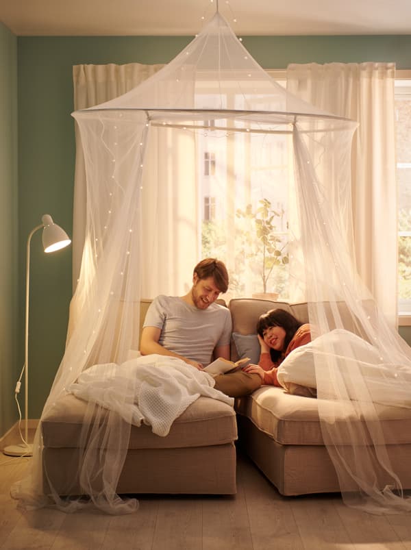 Pár leží pod prikrývkami VÅRELD na dvoch ležadlách umiestnených vedľa seba, slnečné okno za nimi a sieť SOLIG použitá ako baldachýn.