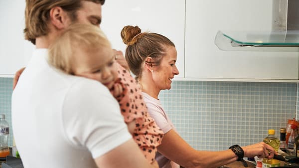 Pár s malým dítětem připravující večeři v kuchyni s bílými dvířky.