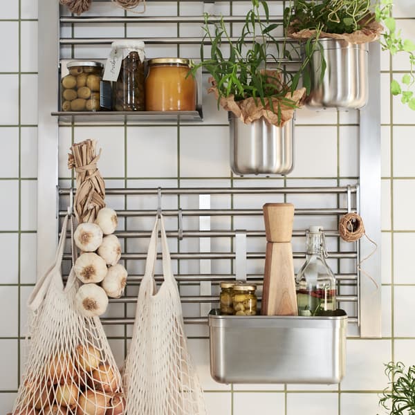Pared de cocina con una rejilla de pared KUNGSFORS y un estante, ganchos y recipientes, todo de acero inoxidable, además de tarros y plantas aromáticas.