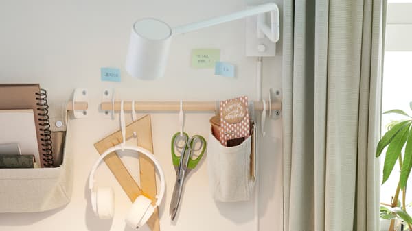 Parete con una lampada da parete NYMÅNE bianca, vari articoli di cancelleria appesi a dei ganci e dentro dei contenitori in canapa NEREBY fissati a un binario in betulla - IKEA