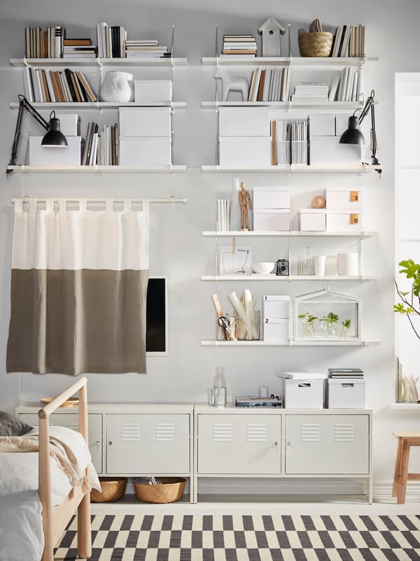 Parete di una camera da letto con mobili IKEA PS e mensole e staffe BERGSHULT/PERSHULT, tutto in bianco. Sulle mensole sono appoggiati contenitori e decorazioni.
