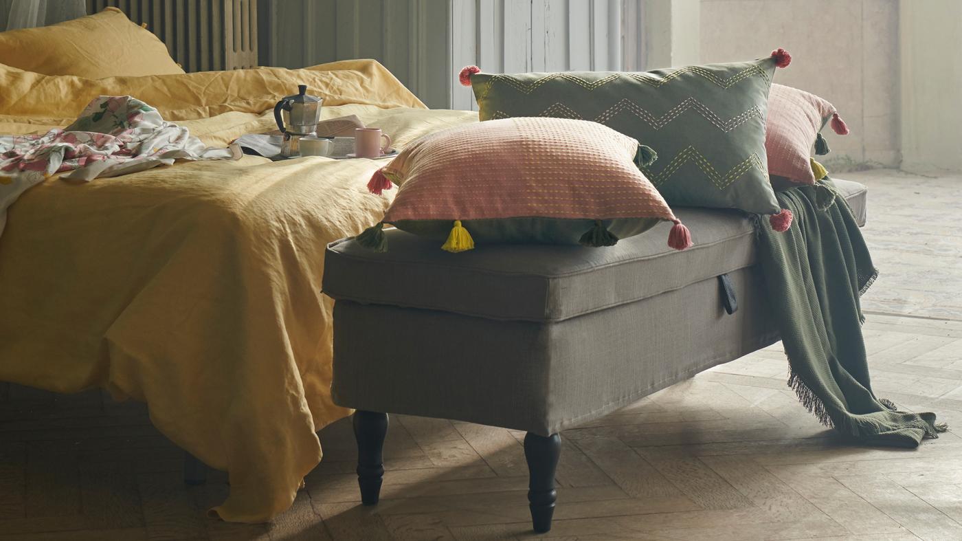 Párnák KLARAFINA és MOAKAJSA huzatban egy STOCKSUND padon fekszenek, egy ágy lábánál, sárga PUDERVIVA ágyneműkkel.