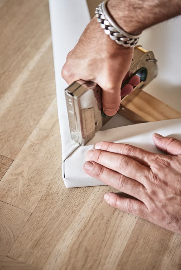 Parquet wooden floor with a person's hands stapling a piece of white canvas onto the corner of a wooden picture frame.