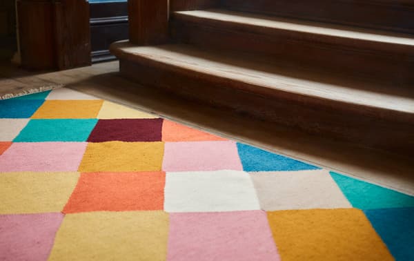 Part of a sunlit VINDERÖD wool rug with a multicoloured, checkered pattern that lies at the base of a staircase.