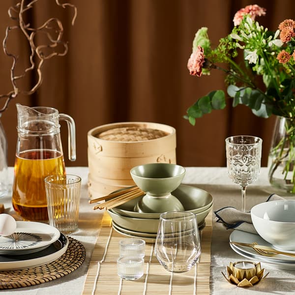 Part of a table with many items on it including flowers, glasses, matt green FÄRGKLAR dishes and a TILLBRINGARE jug.