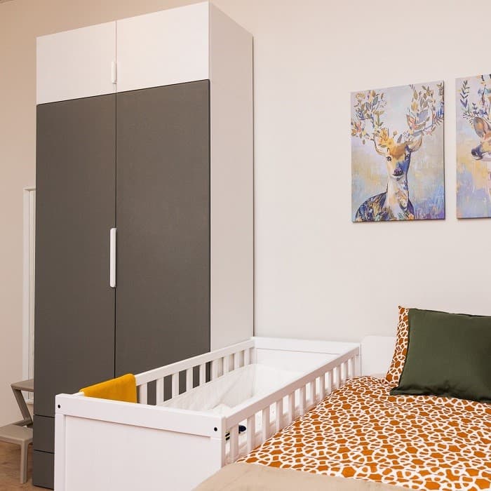 Part of the room with a wardrobe, a baby cot and a bed covered with an orange patterned cover.