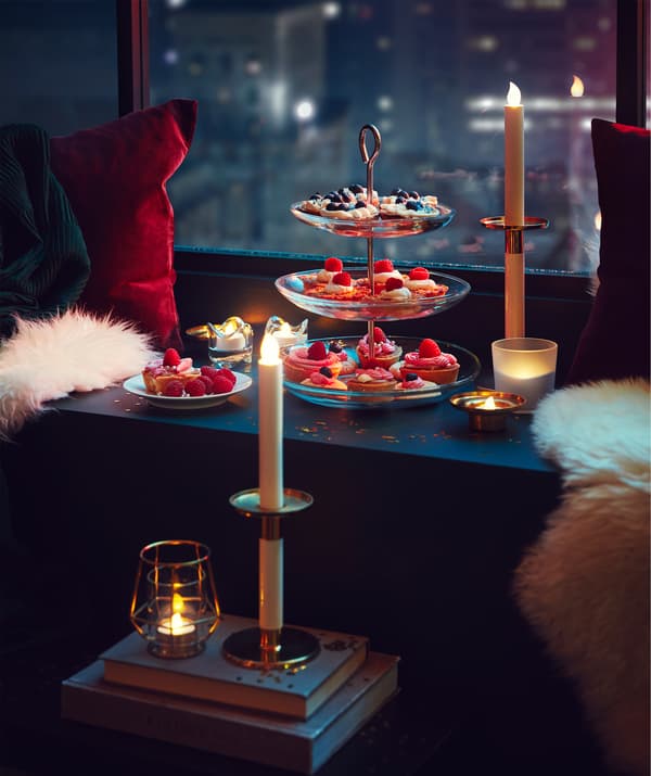 Parte de uma janela, através da qual se vê uma cidade, com um parapeito repleto de iguarias num prato de 3 andares, uma zona acolhedora para sentar e velas LED.