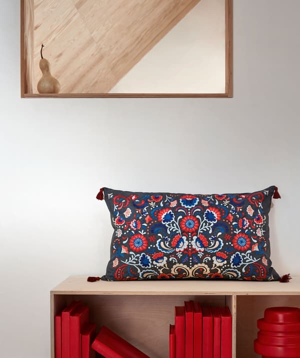 Parte de uma parede com um espelho pendurado e uma estante em baixo com uma almofada bordada colorida colocada em cima.