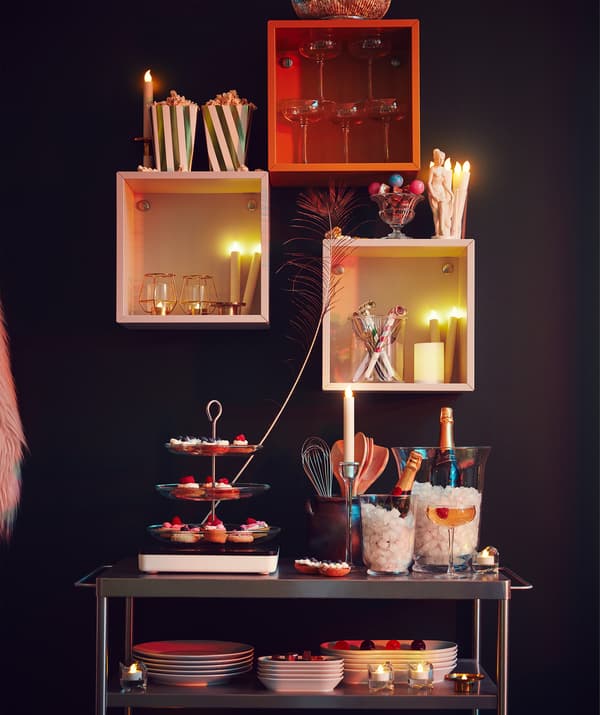 Parte de uma parede com várias estantes em forma de cubos e um carrinho de cozinha por baixo, ambos repletos de acessórios festivos.