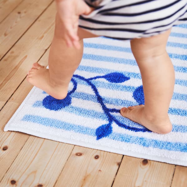 Particolare di un bambino che cammina sul tappeto GULSPARV con le sue strisce blu e bianche e la pianta di mirtillo in un angolo.