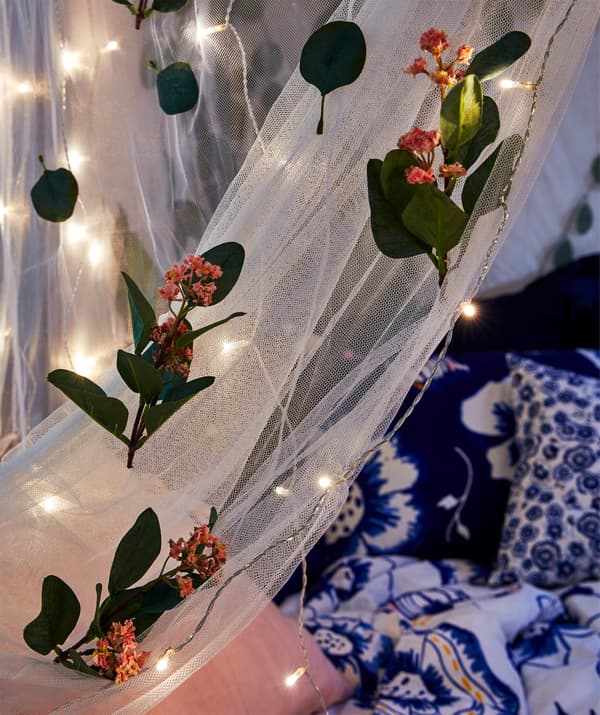 Partie de moustiquaire drapée sur un lit la nuit, décorée de fleurs artificielles et de guirlandes lumineuses.