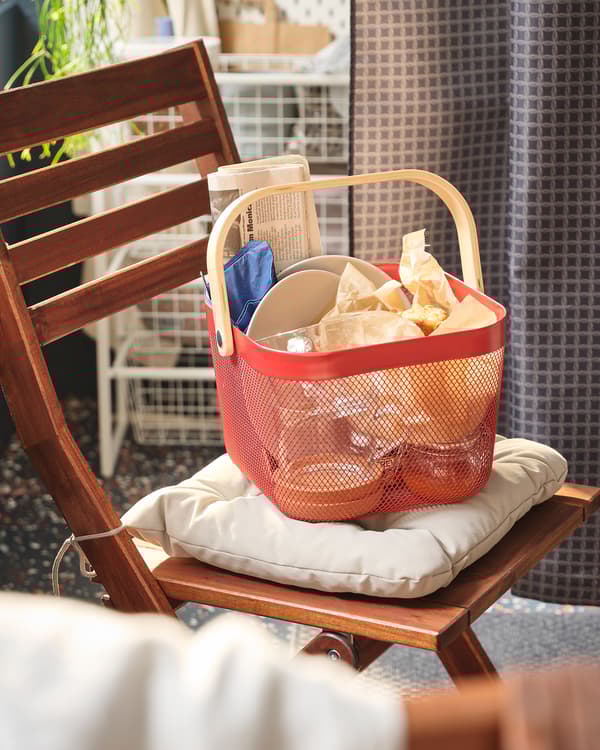 Partie d’un balcon avec un panier rouge RISATORP dans lequel se trouve un petit repas, le tout placé sur le coussin d’une chaise ÄPPLARÖ.