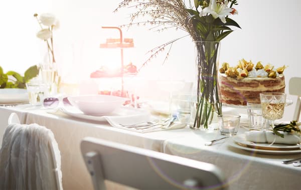 Partie d’une grande table somptueusement dressée de manière romantique avec des meubles, une nappe et de la vaisselle blanche; un gâteau, un plateau avec des cookies et des fleurs.