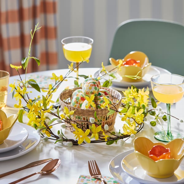 Påskdukning med ett dekorerat bord med porslin och munblåst glas och gula blommor, korg med färglagda ägg i mitten, och glas med gul dryck.