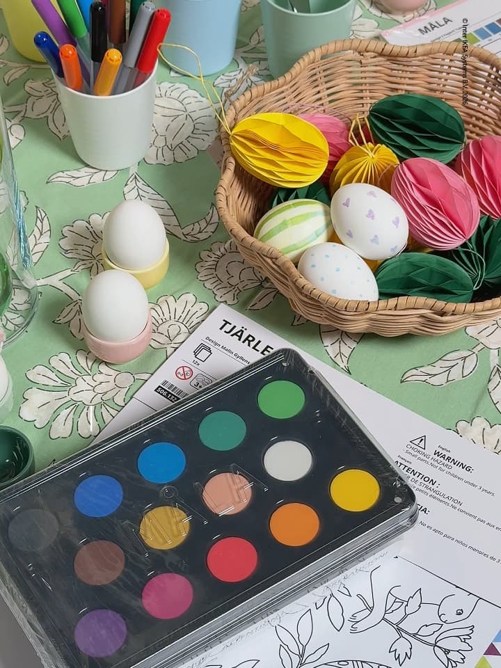 Påskpyssel med allt som behövs för att måla ägg. Vattenfärger, ägg i äggkoppar och färgglada pappersdekorationer i en korg.