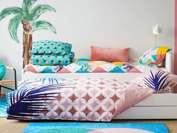 パステルカラーの寝具を使った明るい子ども部屋。ヤシの木柄の壁とカラフルなクッションが特徴。