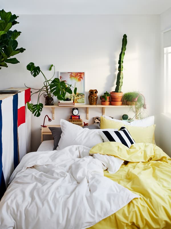 Pat NEIDEN cu două seturi de lenjerie de pat ÄNGSLILJA, unul galben și unul alb. Plante și obiecte decorative pe o poliță deasupra patului.