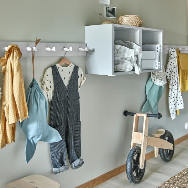 Patères murales blanches avec crochets où les vêtements d’enfants pendent, étagères gris clair et bicyclette pour enfants en bois.