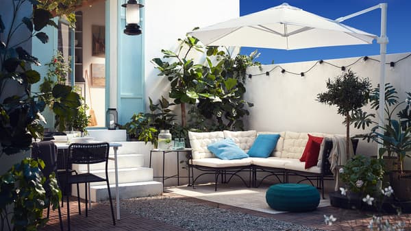 Patio luar yang mempunyai sofa bermodul dengan kusyen berwarna putih, payung berwarna putih, kusyen duduk berwarna biru hijau dan banyak jenis tumbuh-tumbuhan.