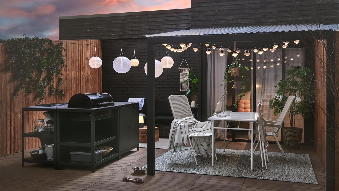 Patio yang disediakan untuk musim bunga dengan pulau dapur dan gril arang GRILLSKÄR, meja dan kerusi TORPARÖ serta lampu.