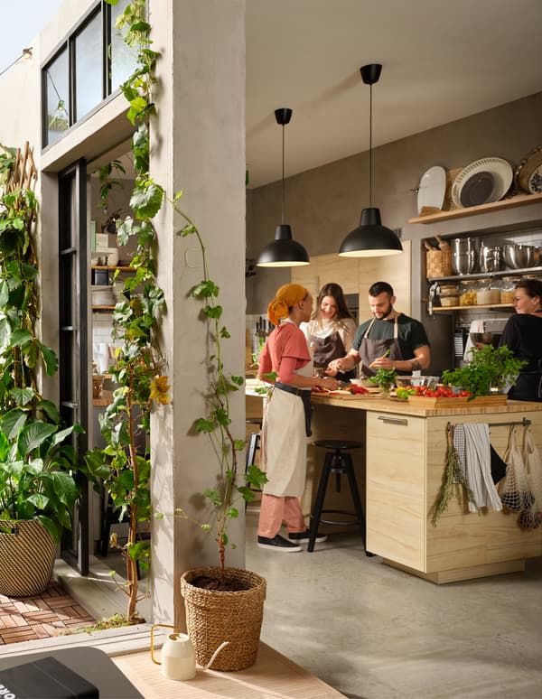 Patru persoane robotesc în jurul unei insule mari de bucătărie, pregătind mâncare. Ușile pe jumătate deschis dau într-o curte cu plante.