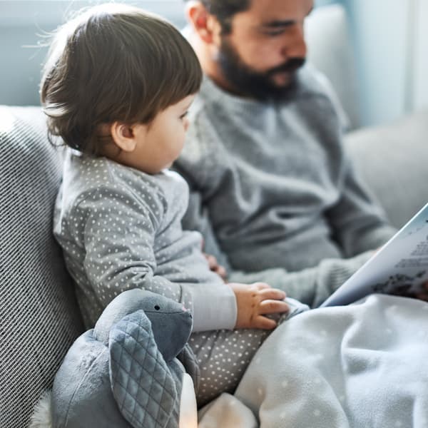 Peluche PEKHULT con luce notturna a LED e una coperta LEN; accanto, un padre che legge al suo bambino su un divano grigio.