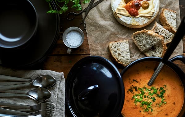 Pentola in acciaio smaltato blu e un mestolo accanto a pane fresco affettato, Camembert al forno, una pila di piatti, tovaglioli e posate.