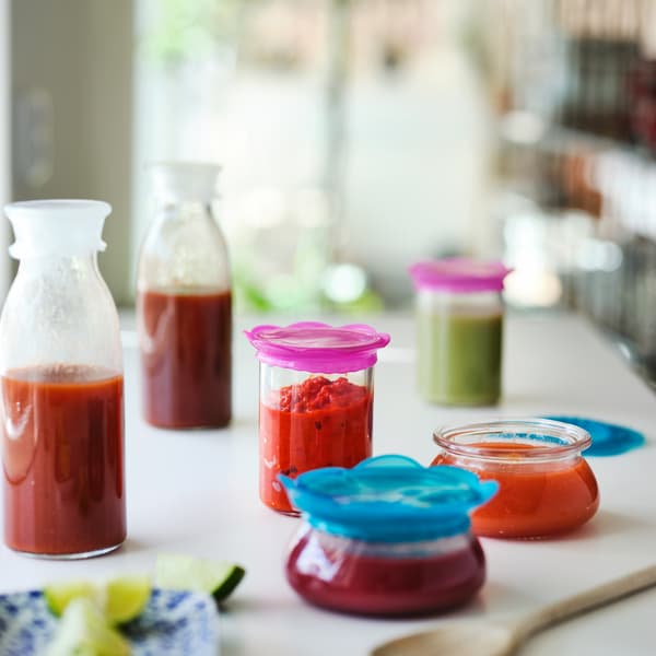 Penutup makanan ÖVERMÄTT silikon merah jambu, lut sinar, berwarna biru firus pada beberapa balang sos, di atas permukaan kerja berwarna putih.