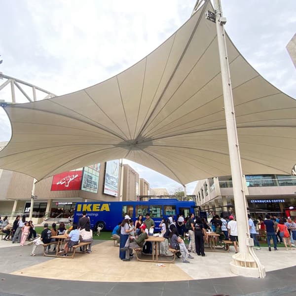 People gathered under a large, white canopy at an outdoor plaza with an IKEA truck and a Starbucks in the background. The atmosphere is lively and social.