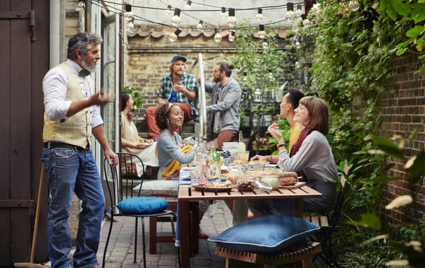 People sitting and standing around a wooden table in a backyard area with brick walls and plants.