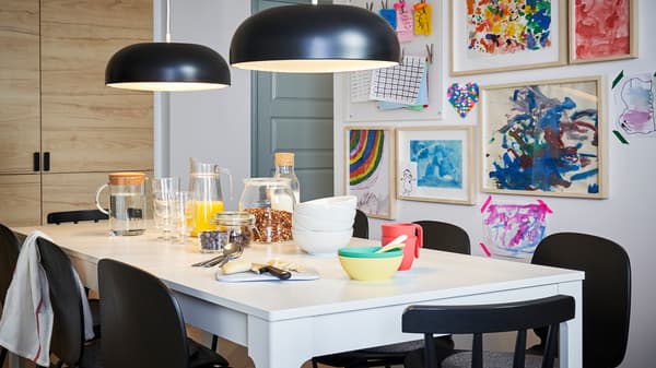 Pequeno-almoço servido numa mesa EKEDALEN em branco com cadeiras em preto e dois candeeiros suspensos em preto, diante de uma parede com desenhos de crianças.
