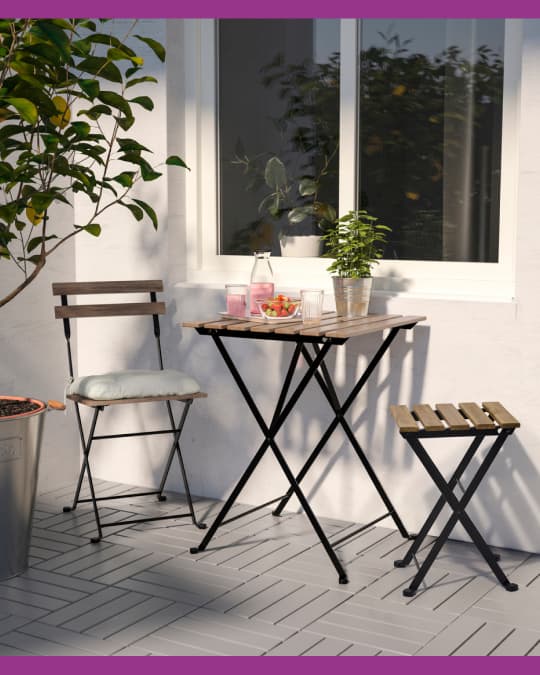 Pequeño balcón con mesa plegable de madera, dos asientos y plantas alrededor, iluminado por luz natural.