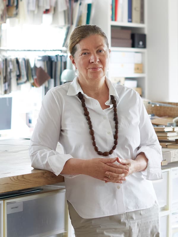 Pereka bentuk Ilse Crawford memakai baju berwarna putih dan kalung yang besar dan tebal sedang bersandar pada permukaan atas kerja di dalam studionya.