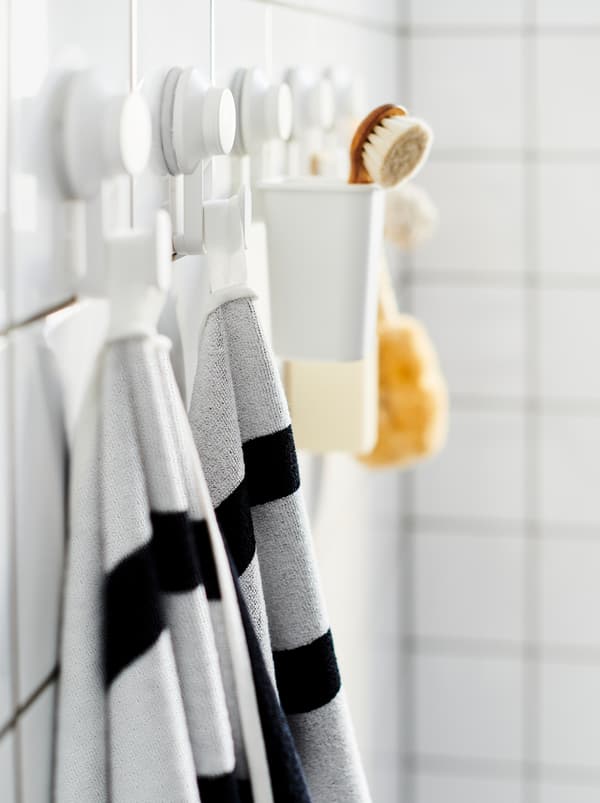 Perete de baie cu faianță albă cu un șir de cârlige cu ventuză TISKEN pe care se află prosoape, un scruber și accesorii similare pentru baie.