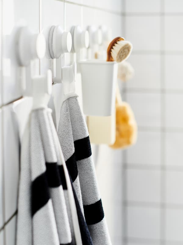 Perete de baie cu faianță cu un șir de cârlige cu ventuză TISKEN pe care se află prosoape, un scruber și accesorii similare.