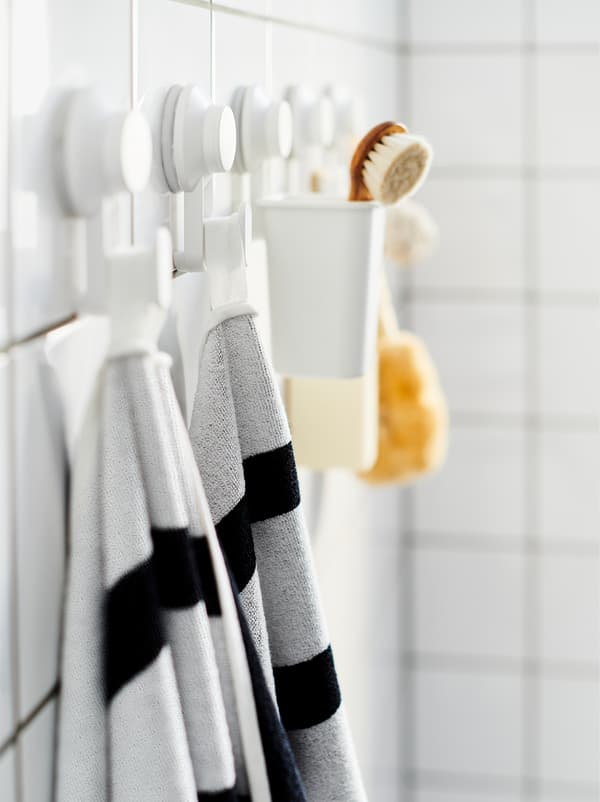 Perete de baie cu faianță cu un șir de cârlige cu ventuză TISKEN pe care se află prosoape, un scruber și accesorii similare pentru baie.