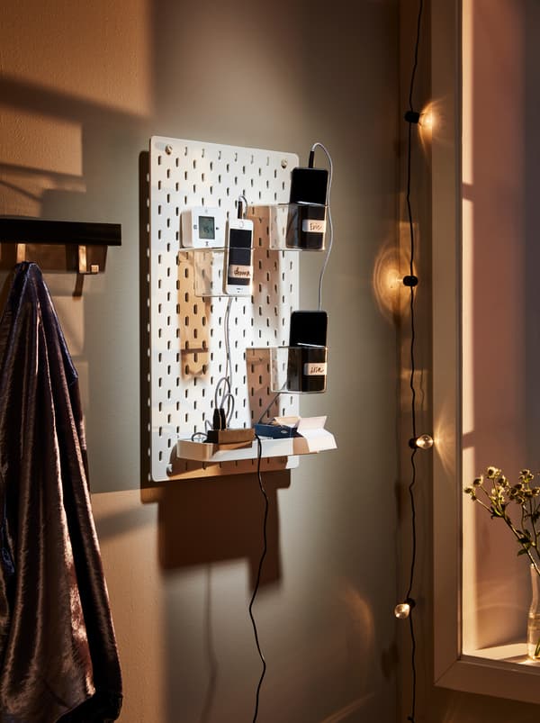 Perete luminat discret pe care este așezat un panou perforat SKÅDIS cu recipiente pentru încărcătoare LÖRBY, telefoane mobile și accesorii pentru telefoane.