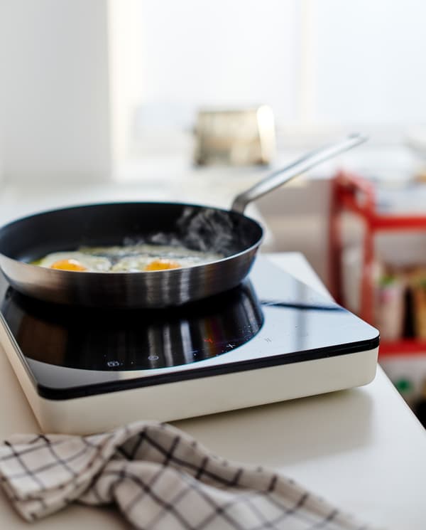 Permukaan atas kerja berwarna putih dengan hob aruhan mudah alih berwarna hitam/putih, tuala pengesat berwarna hitam/putih dan kuali leper yang terdapat telur yang sedang digoreng.