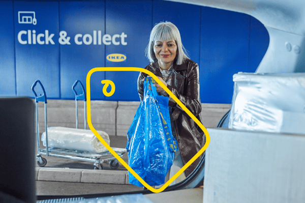 Persoană ridică o pungă albastră IKEA din portbagaj, în zona „Click & collect” a magazinului IKEA; în fundal, două cărucioare și perete albastru cu logo IKEA.