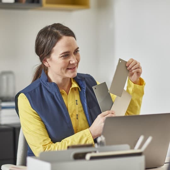 Person am Schreibtisch mit Laptop, hält drei Farbmuster hoch, Büro im Hintergrund.