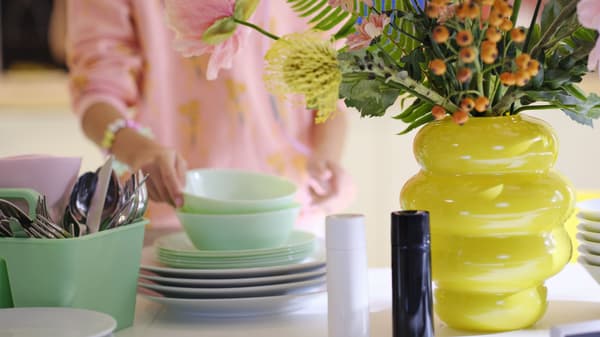 Person arranging pastel dishes on a table beside a bright yellow vase with flowers and a utensil holder.