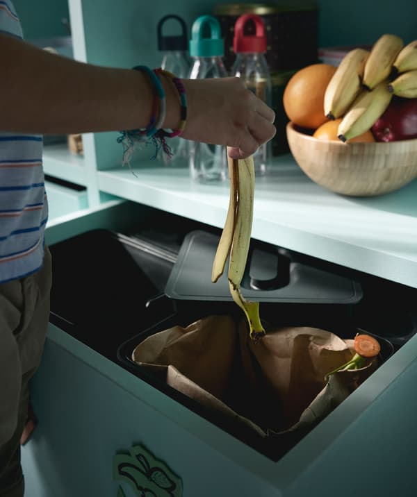Person dropping a banana peel into a black VARIERA recycling bin.