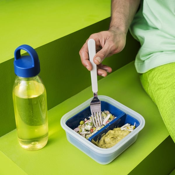 Person eating from a small lunch container beside a water bottle on a bright green surface.