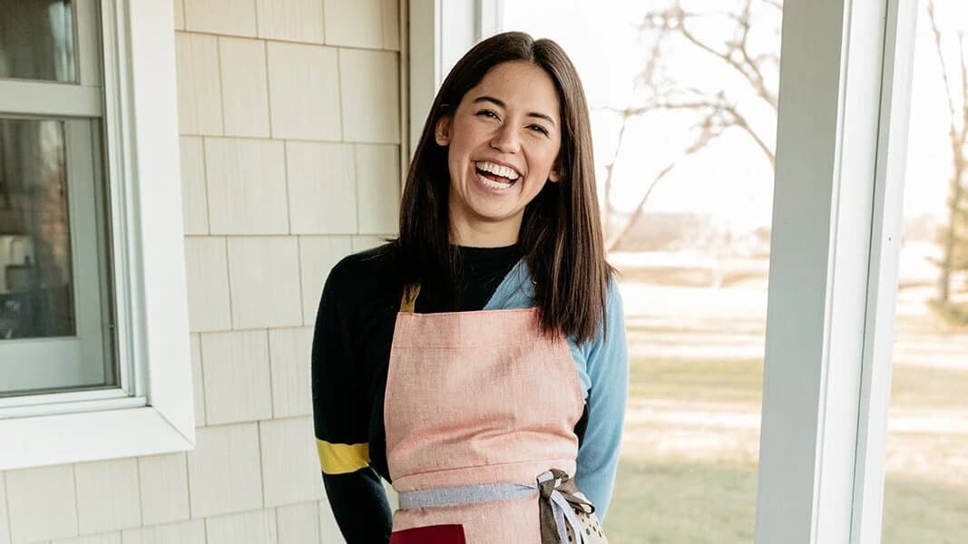 Person facing camera on a porch, wearing a pink apron, standing near a window with a yard behind.