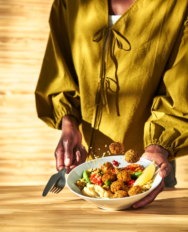 Person hält eine weiße Schüssel mit Falafelbällchen, Hummus, Gemüse und Zitronenspalte. Einige Falafelbällchen schweben über der Schüssel. Hintergrund aus hellem Holz.