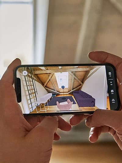 Person hält Smartphone und fotografiert ein gemütlich eingerichtetes Zimmer mit Holzbalkendecke und Tageslicht – Fokus auf digitalem Einrichten und Wohnideen festhalten.