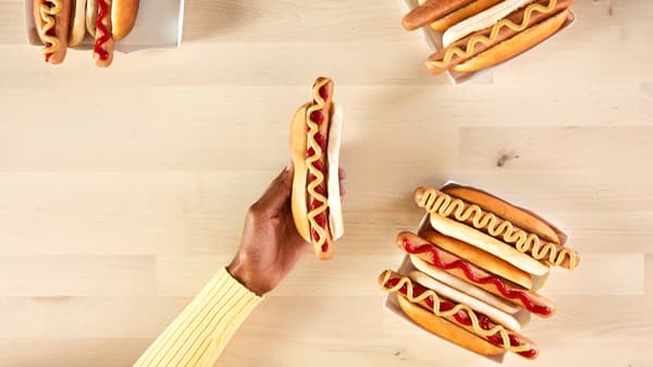 Person håller en växtbaserad korv i bröd med senap, omgiven av sju andra växtbaserade korvar i bröd med ketchup eller senap på ett ljust träbord.


