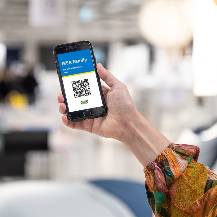 Person holding a smartphone displaying an IKEA Family digital card with a QR code, Lithuanian email address, and the label 'Lietuva', in a bright IKEA store environment.