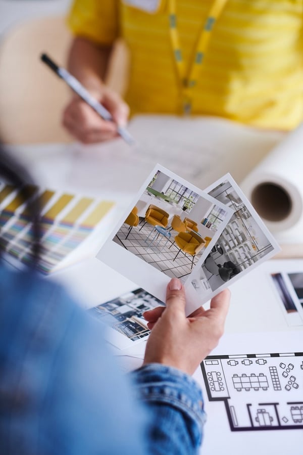 Person holding interior design photos while planning IKEA-style workspace layout with color swatches and floor plan.