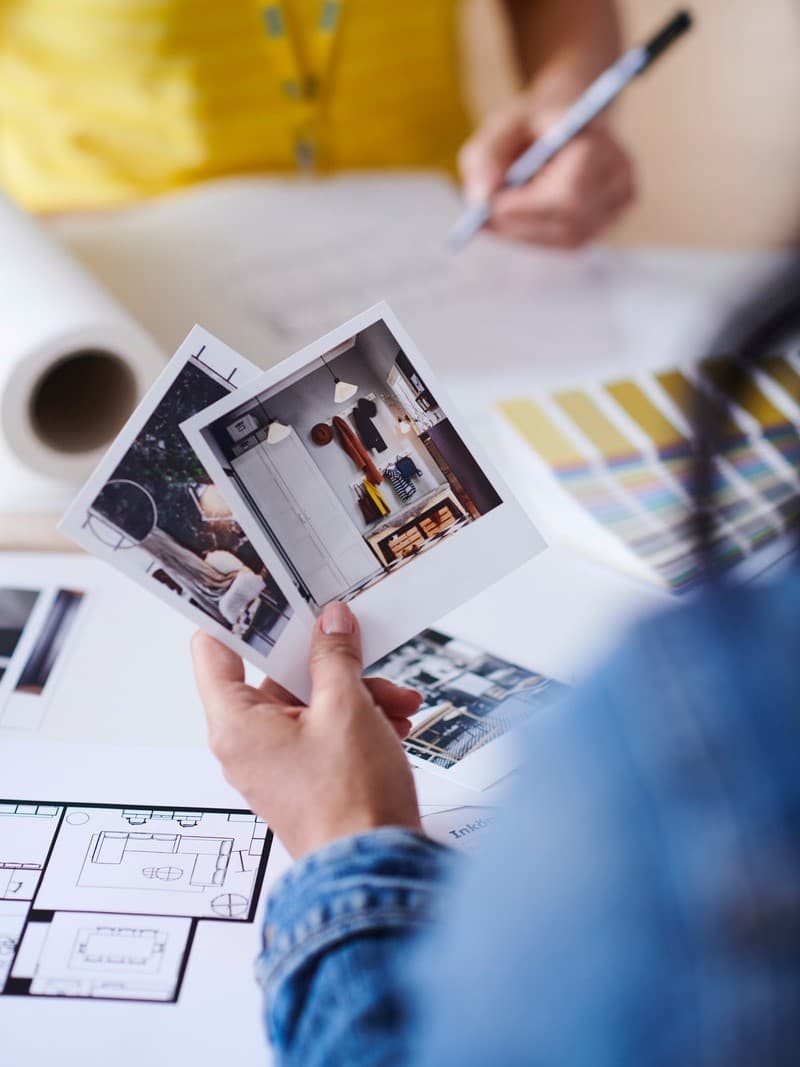 Person holding two photos, one showing an interior; in the background, color swatches and plans on a table.